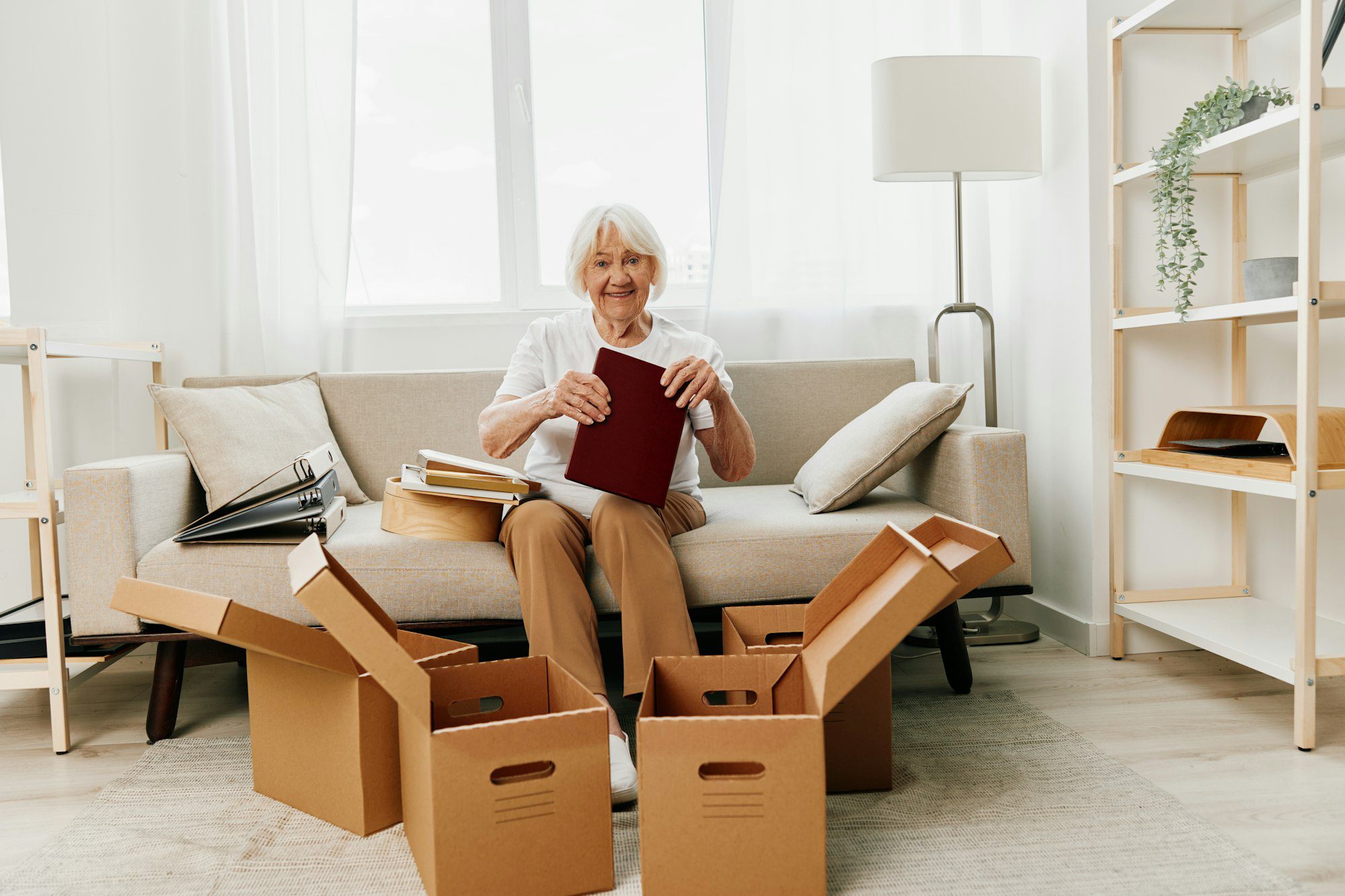 Elderly woman sits on a sofa at home with boxes. collecting things with memories.