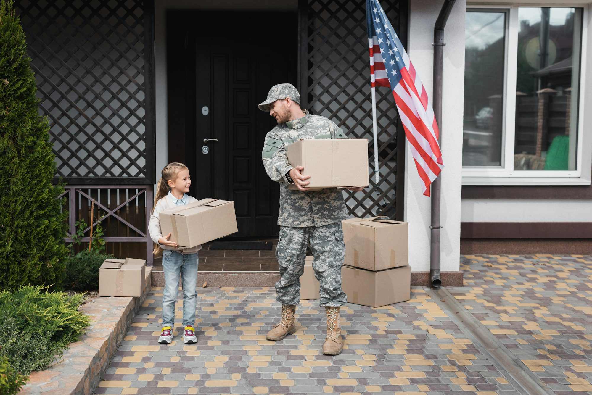 Father in military uniform and daughter holding cardboard boxes near house with american flag